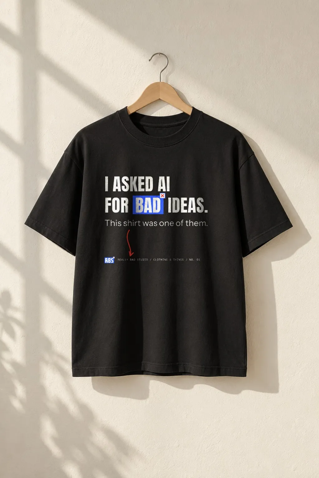 The bad ideas tee, black, hanging on a wooden hanger against a sunlit wall.
