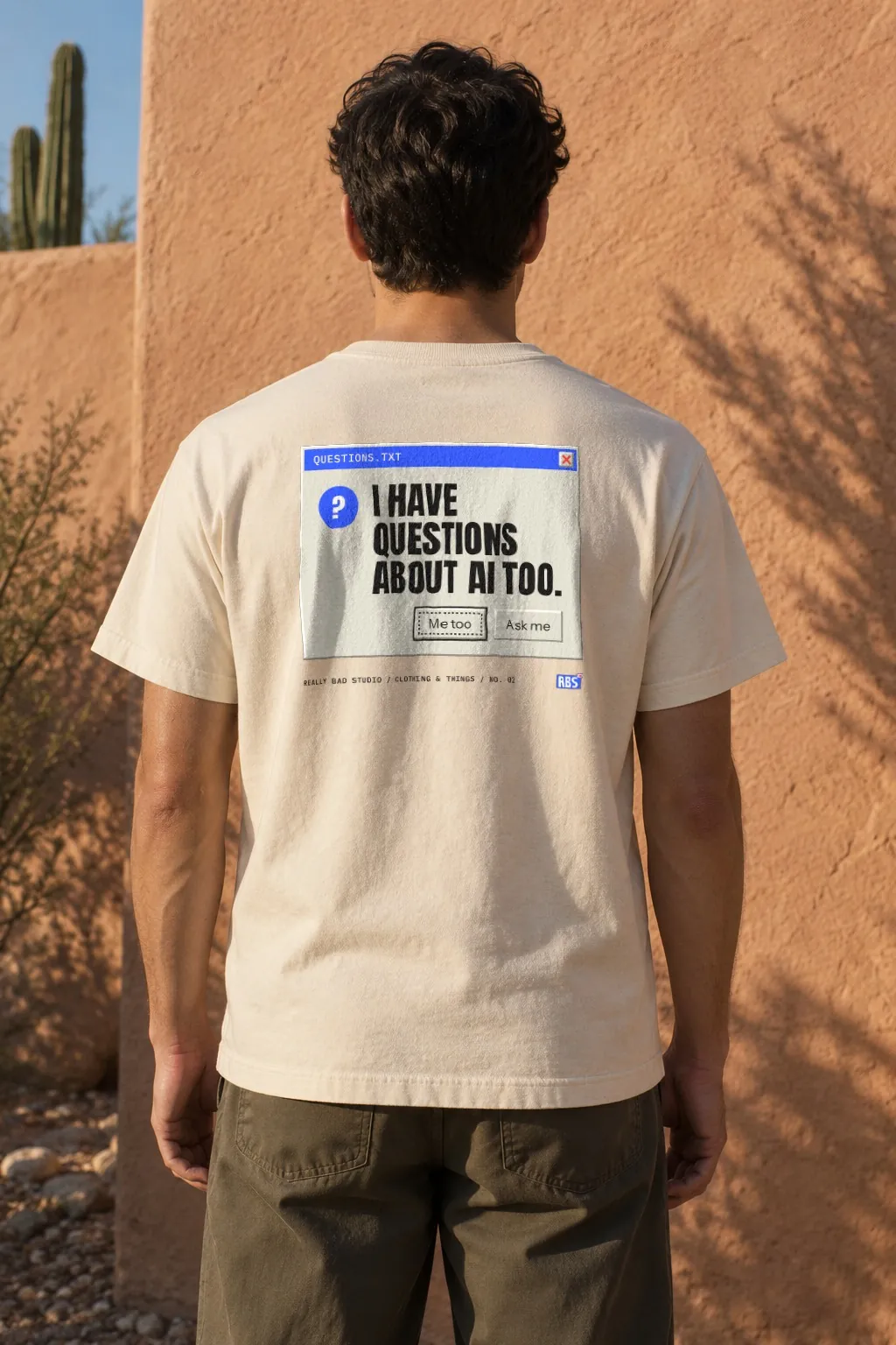 Seen from behind, a person in a natural tee with the questions window printed across the back, facing an adobe wall.
